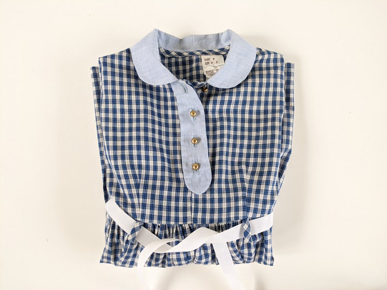 Op de afbeelding: Een blauw-wit geruit jurkje met een witte kraag en een witte strik. Het jurkje heeft een knoopsluiting en een label met de tekst "SIZE 4, LEFT P, STYLE 1".