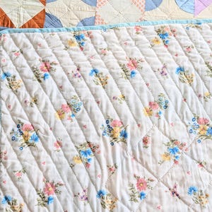 Puede incluir: Primer plano de una colcha blanca acolchada con un estampado floral en rosa, azul y amarillo. La colcha tiene un borde azul claro y un patr&oacute;n cosido en diagonal. La colcha forma parte de una colecci&oacute;n m&aacute;s grande de colchas hechas a mano.