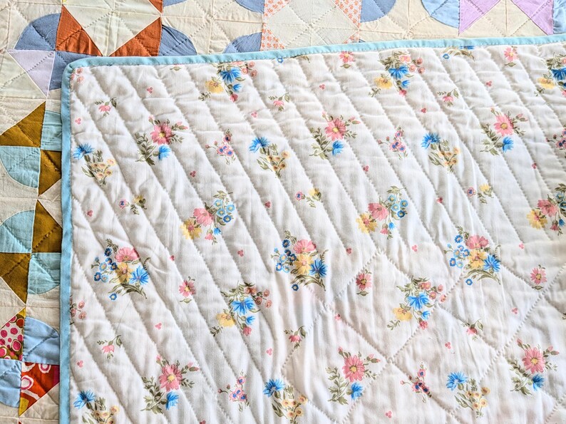 Puede incluir: Primer plano de una colcha blanca acolchada con un estampado floral en rosa, azul y amarillo. La colcha tiene un borde azul claro y un patr&oacute;n cosido en diagonal. La colcha forma parte de una colecci&oacute;n m&aacute;s grande de colchas hechas a mano.