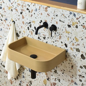 Op de afbeelding: Een beige keramiek badkamer wastafel met een afgeronde rand en een zwarte afvoer. De wastafel is gemonteerd aan een muur met zwarte kranen en een witte en bruine terrazzo tegel achtergrond.