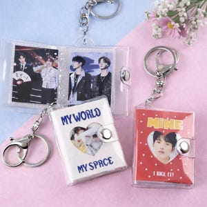 Peut inclure: Porte-clés avec albums photo en plastique transparent et anneaux argentés. Un porte-clés a une couverture blanche avec le texte "MY WORLD MY SPACE". Un autre a une couverture rouge avec le texte "MINE I LIKE IT!". Les albums contiennent des photos de personnes.