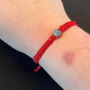 May include: A red string bracelet with a grey stone bead. The bracelet is tied around a person's wrist.