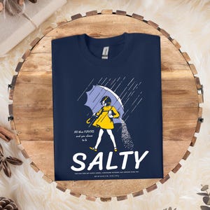 Puede incluir: Camiseta azul marino con un gr&aacute;fico de una persona bajo un paraguas, con la palabra "SALTY" en blanco. El gr&aacute;fico incluye el texto "All these flavors and you choose to be".