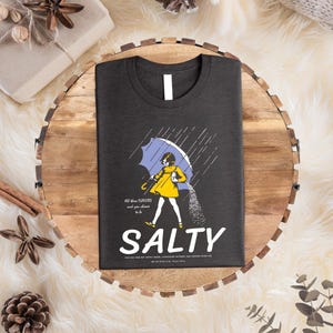Puede incluir: Camiseta gris oscuro con un gr&aacute;fico de una persona bajo un paraguas azul, vertiendo sal. La palabra "SALTY" est&aacute; debajo del gr&aacute;fico, con el texto "All these flavors and you choose to be" encima. La camiseta est&aacute; sobre una bandeja de madera.