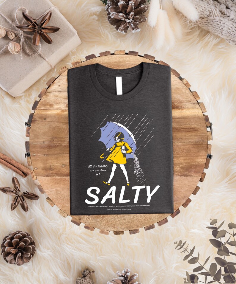Puede incluir: Camiseta gris oscuro con un gr&aacute;fico de una persona bajo un paraguas azul, vertiendo sal. La palabra "SALTY" est&aacute; debajo del gr&aacute;fico, con el texto "All these flavors and you choose to be" encima. La camiseta est&aacute; sobre una bandeja de madera.