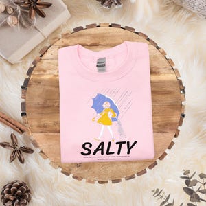 Puede incluir: Sudadera rosa claro con un gr&aacute;fico de una persona con un vestido amarillo y un paraguas azul bajo la lluvia. La palabra "SALTY" est&aacute; impresa en negro debajo. La sudadera est&aacute; doblada sobre una superficie de madera.