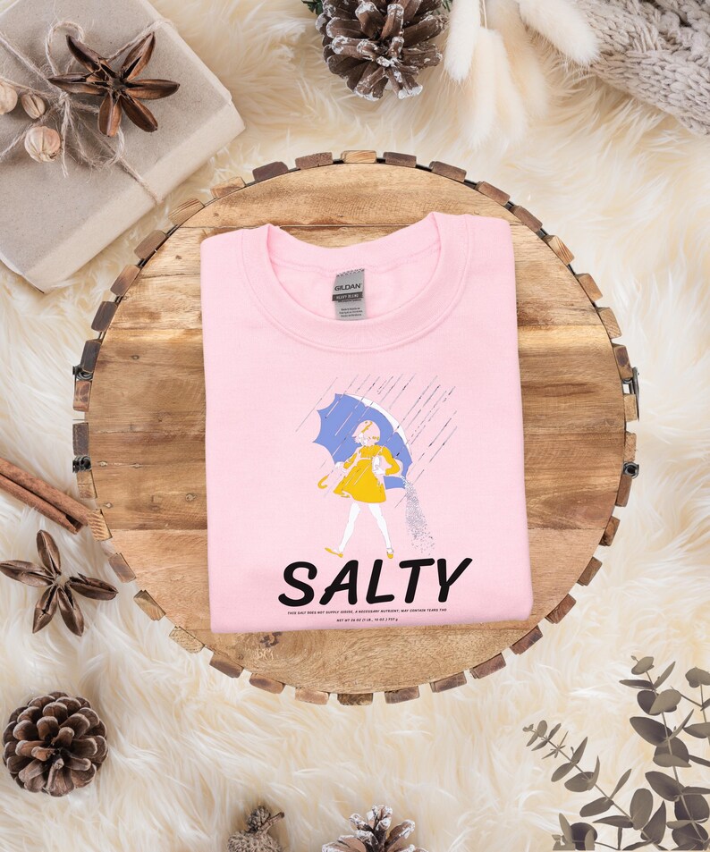 Puede incluir: Sudadera rosa claro con un gr&aacute;fico de una persona con un vestido amarillo y un paraguas azul bajo la lluvia. La palabra "SALTY" est&aacute; impresa en negro debajo. La sudadera est&aacute; doblada sobre una superficie de madera.