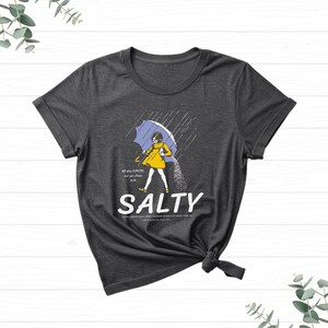 Puede incluir: Camiseta gris oscuro con una ilustraci&oacute;n humor&iacute;stica. Una persona con un impermeable amarillo, sosteniendo un paraguas azul, vierte sal bajo la lluvia. La palabra "SALTY" est&aacute; debajo, con texto adicional arriba y abajo.