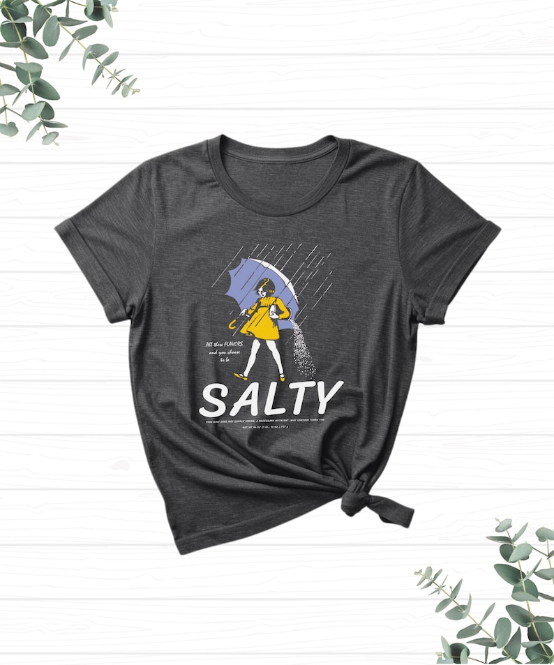 Puede incluir: Camiseta gris oscuro con una ilustraci&oacute;n humor&iacute;stica. Una persona con un impermeable amarillo, sosteniendo un paraguas azul, vierte sal bajo la lluvia. La palabra "SALTY" est&aacute; debajo, con texto adicional arriba y abajo.