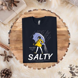 Puede incluir: Camiseta azul marino con un gr&aacute;fico de una persona bajo un paraguas, con la palabra "SALTY" en letras blancas grandes. La imagen incluye el texto "All these flavors and you choose to be".