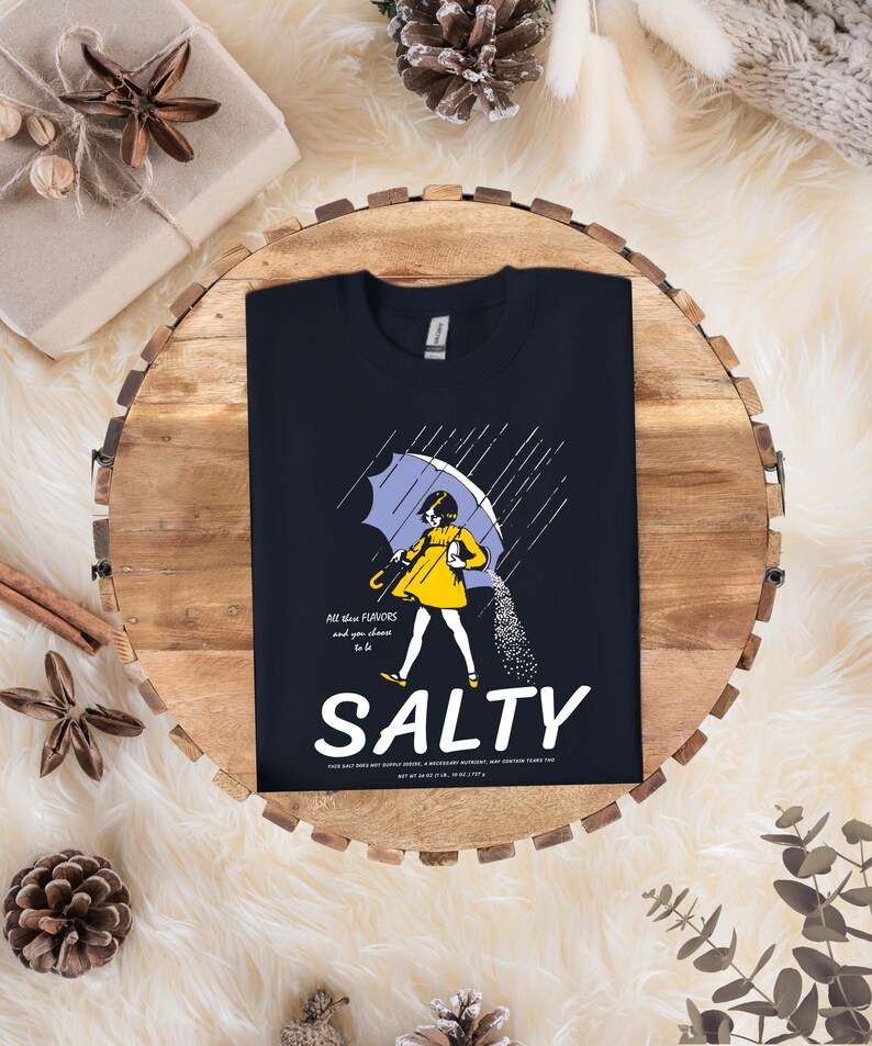 Puede incluir: Camiseta azul marino con un gr&aacute;fico de una persona bajo un paraguas, con la palabra "SALTY" en letras blancas grandes. La imagen incluye el texto "All these flavors and you choose to be".