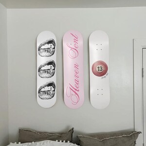 May include: Three skateboard decks mounted on a wall. One white deck features a black and white illustration of teeth. A pink deck has the words "Heaven Sent" in cursive. A white deck displays a red 13-ball.