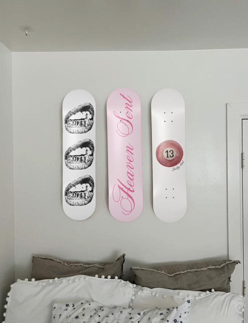 May include: Three skateboard decks mounted on a wall. One white deck features a black and white illustration of teeth. A pink deck has the words "Heaven Sent" in cursive. A white deck displays a red 13-ball.