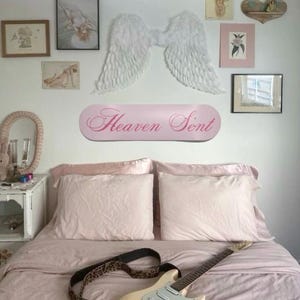 May include: A bedroom scene with a light pink bedspread and pillows. A cream-colored electric guitar with a leopard print strap rests on the bed. Above the bed, a pink sign reads "Heaven Sent", and decorative angel wings are mounted on the wall. Various framed artworks complete the decor.
