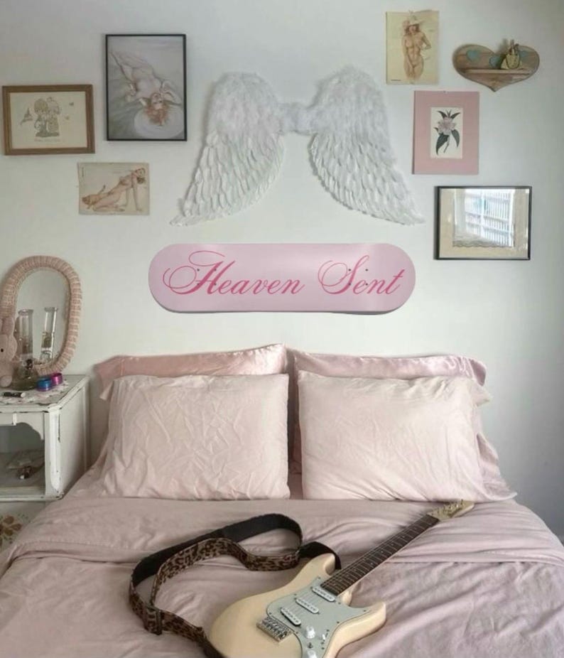 May include: A bedroom scene with a light pink bedspread and pillows. A cream-colored electric guitar with a leopard print strap rests on the bed. Above the bed, a pink sign reads "Heaven Sent", and decorative angel wings are mounted on the wall. Various framed artworks complete the decor.