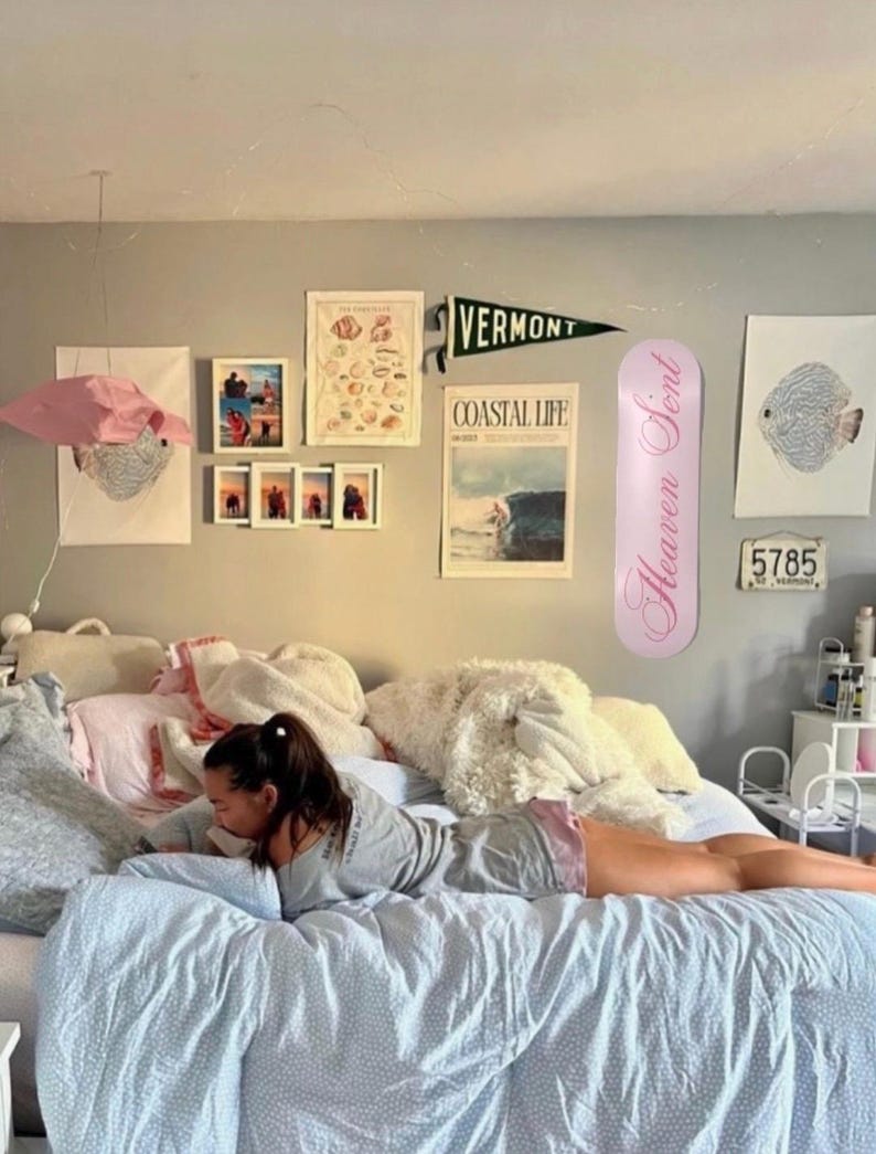 May include: A bedroom scene with a bed covered in pillows and a blue patterned comforter. Wall decor includes framed art, a Vermont pennant, and a pink decorative item. A pink surfboard-shaped item with the text "Heaven Scent" is leaning against the wall.