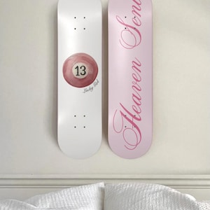 May include: Two skateboards mounted on a wall. One is white with a pool ball graphic and the number 13. The other is pink with the words "Heaven Sent" in cursive. The skateboards are above a white bed.