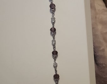 May include: A beaded accessory featuring alternating clear and purple beads strung on a silver-colored chain. The accessory has a lobster clasp at the top and a decorative bead at the bottom. The beads are faceted, reflecting light.