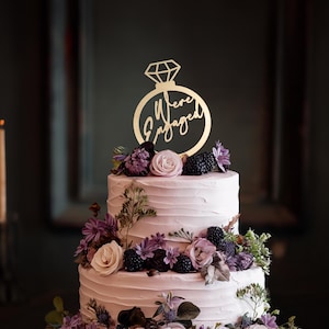 May include: A two-tiered wedding cake with light pink frosting and floral decorations. The cake is topped with a gold ring-shaped cake topper that reads "We're Engaged". The flowers include roses, blackberries, and other purple and green foliage.