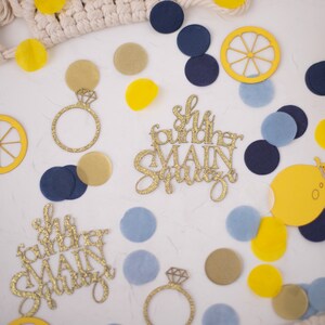 May include: A collection of party decorations, including gold glitter ring cutouts, lemon slices, and confetti in yellow, navy, and light blue. The phrase "She found her MAIN SQUEEZE" is displayed in gold glitter script. The items are scattered on a white surface.