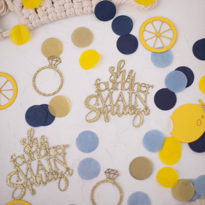 May include: A collection of party decorations, including gold glitter ring cutouts, lemon slices, and confetti in yellow, navy, and light blue. The phrase "She found her MAIN SQUEEZE" is displayed in gold glitter script. The items are scattered on a white surface.