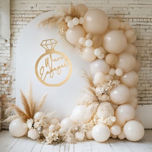 May include: A white backdrop with a gold ring and the text "We're Engaged" in a script font. The backdrop is decorated with a garland of cream and beige balloons and dried pampas grass.