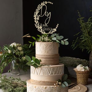 Könnte beinhalten: Eine dreistöckige Hochzeitstorte mit einem Holzaufsatz. Die Torte ist mit weißem Zuckerguss, Jutegarn und Spitzendetails verziert. Der Aufsatz lautet "D & J, it was always you" in Schreibschrift, umgeben von floralen Akzenten.