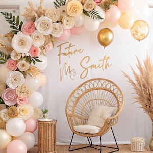 May include: A white backdrop with a floral arch made of paper flowers and balloons. The arch is decorated with greenery and pampas grass. The text "Future Mrs. Smith" is written in gold script on the backdrop. A wicker chair with a white cushion sits in front of the arch.