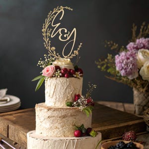 Może przedstawiać: Dwupiętrowy tort weselny z drewnianym topperem z literami "E&J" otoczonymi kwiatowym wieńcem. Ciasto jest udekorowane świeżymi kwiatami i jagodami. Bukiet kwiatów znajduje się w tle.