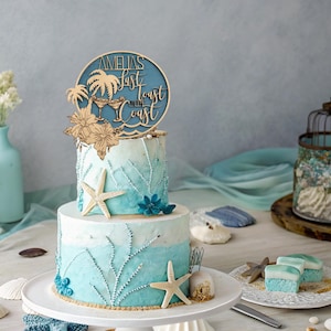 May include: A two-tiered cake decorated with a blue and white ombre frosting, seashells, and starfish. The cake is topped with a wooden sign that reads "Amelia's Last Toast on the Coast".