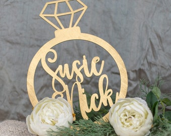 May include: Gold ring cake topper with a diamond design and the names Susie & Nicky written inside the ring.