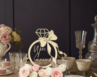 Puede incluir: Decoración de anillo de compromiso de madera con las palabras "We're Engaged" recortadas. El anillo presenta una parte superior en forma de diamante y un lazo. La decoración está rodeada de flores, cristalería y un pastel.