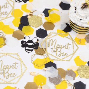 May include: Gold glitter confetti with the text "Meant to Bee" in a geometric frame. The confetti is scattered on a white surface with black and yellow honeycomb shapes, and a cupcake with a bee on top.