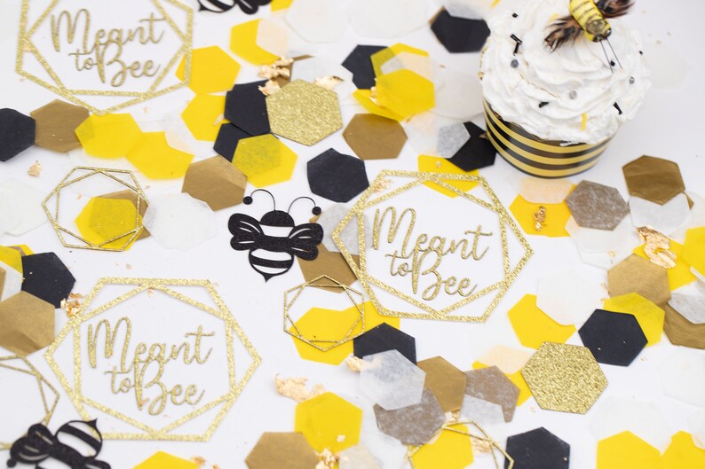 May include: Gold glitter confetti with the text "Meant to Bee" in a geometric frame. The confetti is scattered on a white surface with black and yellow honeycomb shapes, and a cupcake with a bee on top.