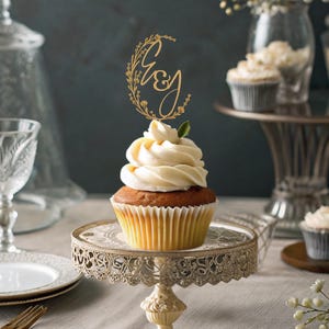 Op de afbeelding: Een gouden cupcake topper met de letters "G & J" omringd door bloemmotieven. De topper is geplaatst op een witte cupcake met vanille frosting.
