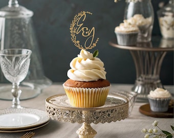 Può includere: Un topper per cupcake dorato con le lettere "G & J" circondate da motivi floreali. Il topper è posizionato su un cupcake bianco con glassa alla vaniglia.