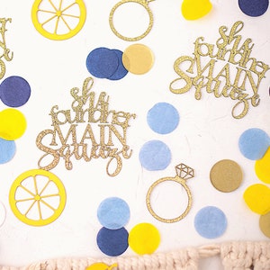 May include: Overhead shot of wedding-themed confetti and decorations. Gold glitter cutouts of a ring and the phrase "She found her MAIN SQUEEZE" are scattered among yellow, blue, and gold confetti pieces. Lemon slice cutouts are also present.