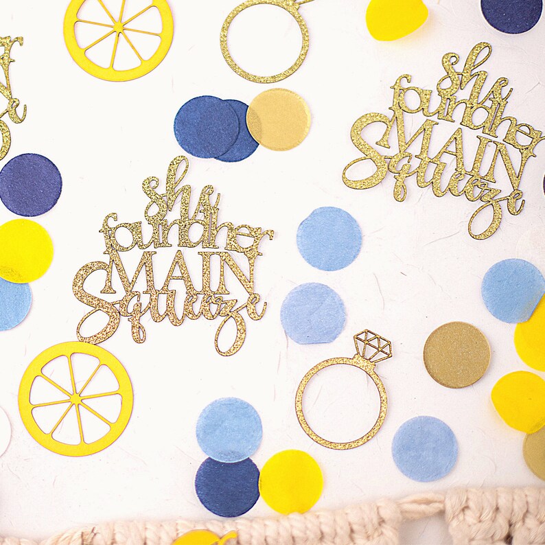 May include: Overhead shot of wedding-themed confetti and decorations. Gold glitter cutouts of a ring and the phrase "She found her MAIN SQUEEZE" are scattered among yellow, blue, and gold confetti pieces. Lemon slice cutouts are also present.