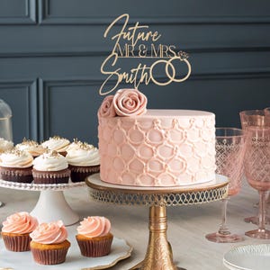 Op de afbeelding: Een roze taart met een delicaat patroon is bedekt met een houten taarttopper waarop "Future Mr & Mrs Smith" staat met twee ringen en een diamant. De taart is omringd door cupcakes en roze glazen.