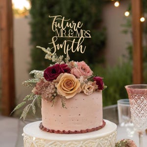 May include: A wedding cake topper with the words "Future Mr & Mrs Smith" in gold script lettering. The topper is placed on a pink cake decorated with burgundy and peach flowers.