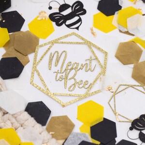 May include: Gold glitter confetti with hexagonal shapes in black, white, and yellow. The confetti is scattered on a white surface with two gold glitter geometric frames that say "Meant to Bee".