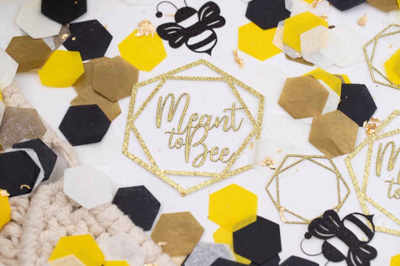 May include: Gold glitter confetti with hexagonal shapes in black, white, and yellow. The confetti is scattered on a white surface with two gold glitter geometric frames that say "Meant to Bee".
