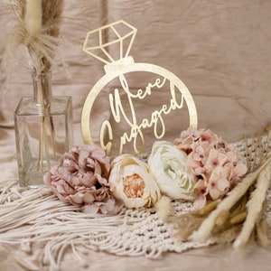 May include: A gold-colored wooden ring-shaped sign with the words "We're Engaged" cut out. The sign is surrounded by pink and cream-colored flowers, dried grasses, and a clear glass bottle. The background is a neutral beige.