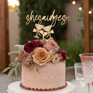 May include: A wooden cake topper with the text "She said yes" and two champagne glasses with bows. The topper is on a pink cake with burgundy and peach flowers.