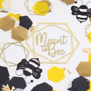 May include: Gold glitter confetti with black and yellow honeycomb shapes, a black bee cutout, and a gold glitter geometric frame with the text "Meant to Bee".