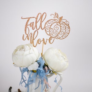 Könnte beinhalten: Ein goldener Glitzer-Kuchenaufsatz mit den Worten "Fall in Love" und einem Kürbis-Design. Der Aufsatz befindet sich in einer weißen Vase mit weißen Blumen und blauen und rosa Papierstreifen.