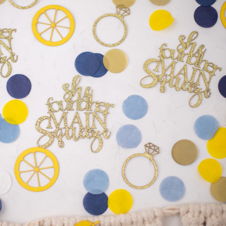 May include: A collection of party decorations, including gold glitter ring cutouts, lemon slices, and confetti in yellow, blue, and gold. The phrase "She found her MAIN Squeeze" is written in gold glitter. Perfect for a bridal shower or engagement party.