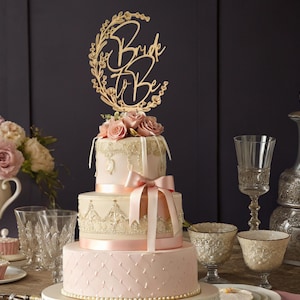 May include: A three-tiered wedding cake with a gold "Bride to Be" cake topper. The cake is decorated with pink frosting, lace details, and a pink ribbon. The top tier is adorned with pink roses.