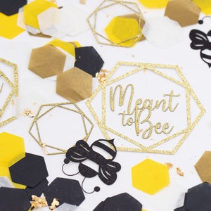 May include: Gold glitter confetti with the text "Meant to Bee" in a geometric frame. There are also black and yellow honeycomb shapes, and black paper cutouts of bees.