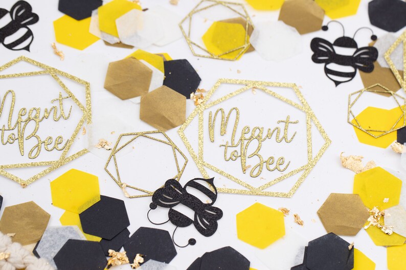 May include: Gold glitter confetti with the text "Meant to Bee" in a geometric frame. There are also black and yellow honeycomb shapes, and black paper cutouts of bees.